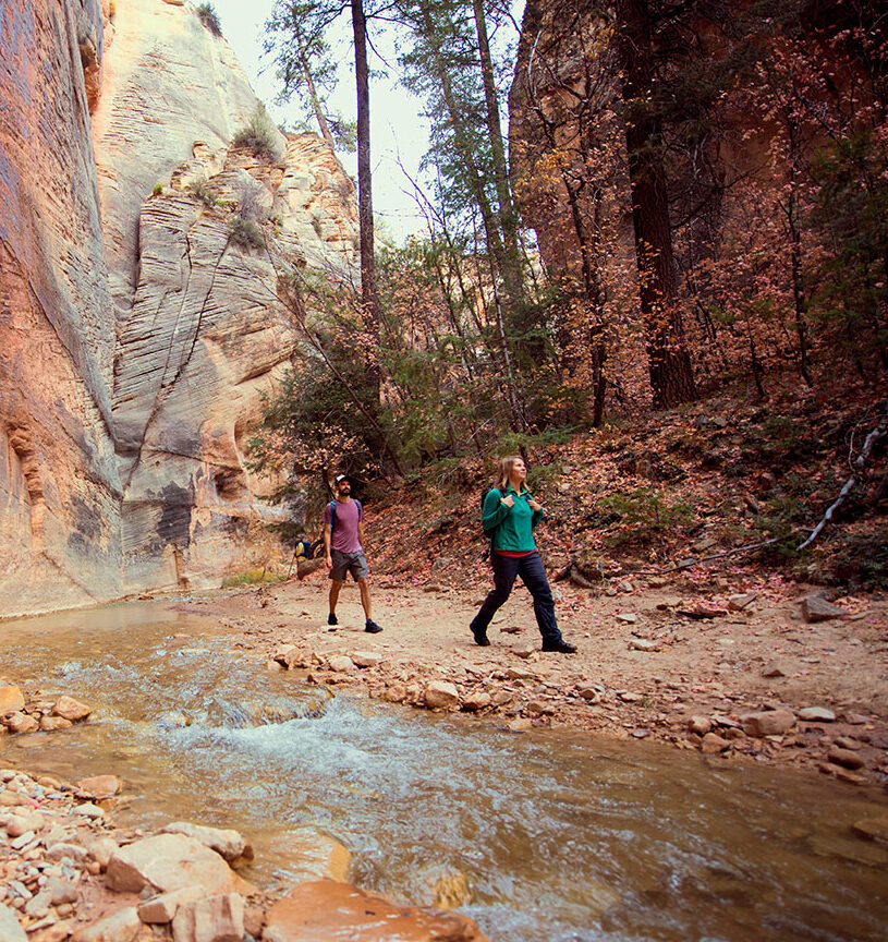 Two hikers walk along a stream in a narrow slot canyon.