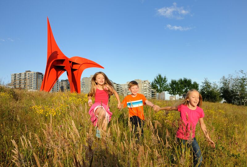 Three children running through tall grass near a red sculpture.