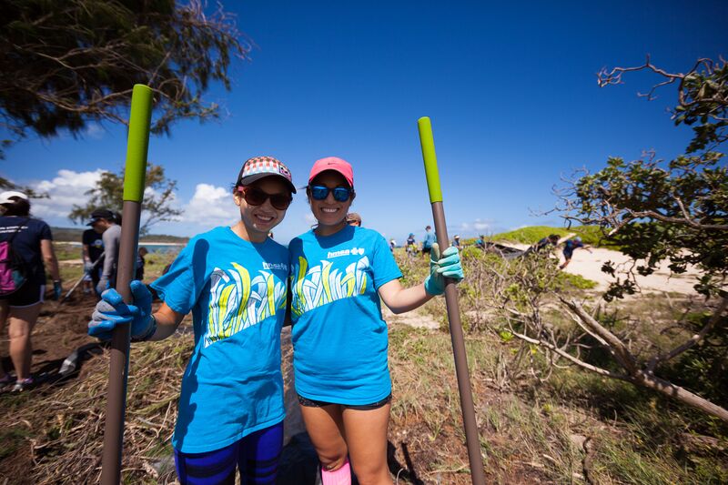 Two women in blue shirts holding sticks on a dirt path.