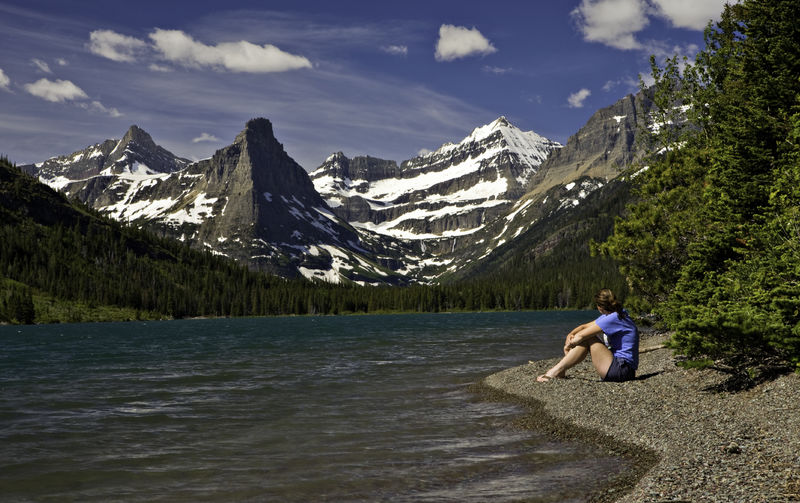 A woman sits on the shore of a lake in Glacier National Park looking towards a mountain.
