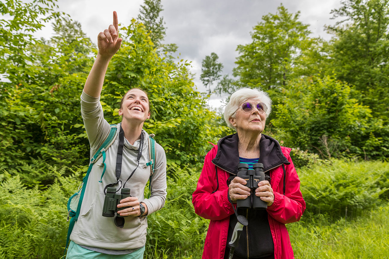 Two women standing with binoculars; one of them is pointing up at something.