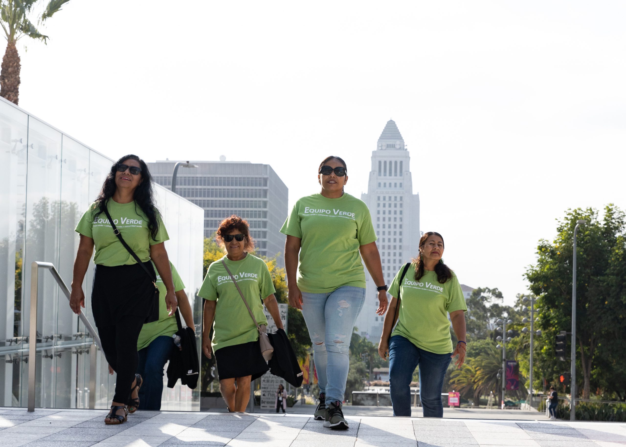 Five women in matching green t-shirts climb the stairs in front of Los Angeles City Hall. 