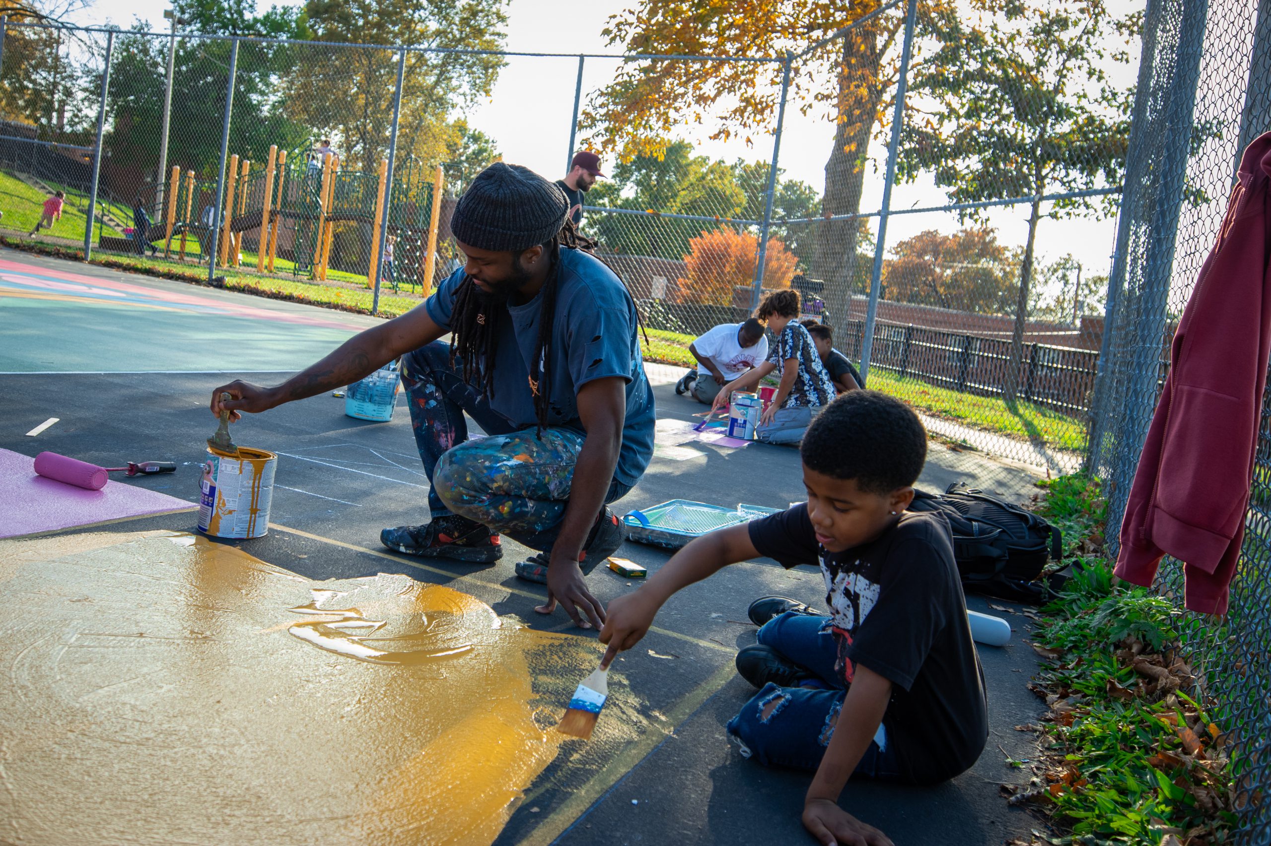 A school-aged boy paints a mural on the surface of a basketball court with help from a young man.