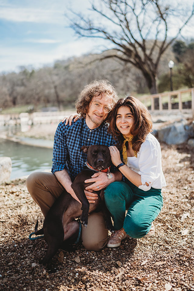 A man and woman posing with their dog in front of a lake.