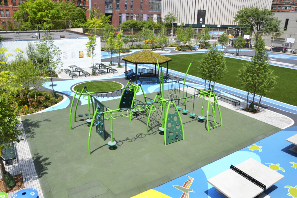 A renovated schoolyard includes inclusive playground equipment and soft, porous surfaces, trees, and a shade structure.