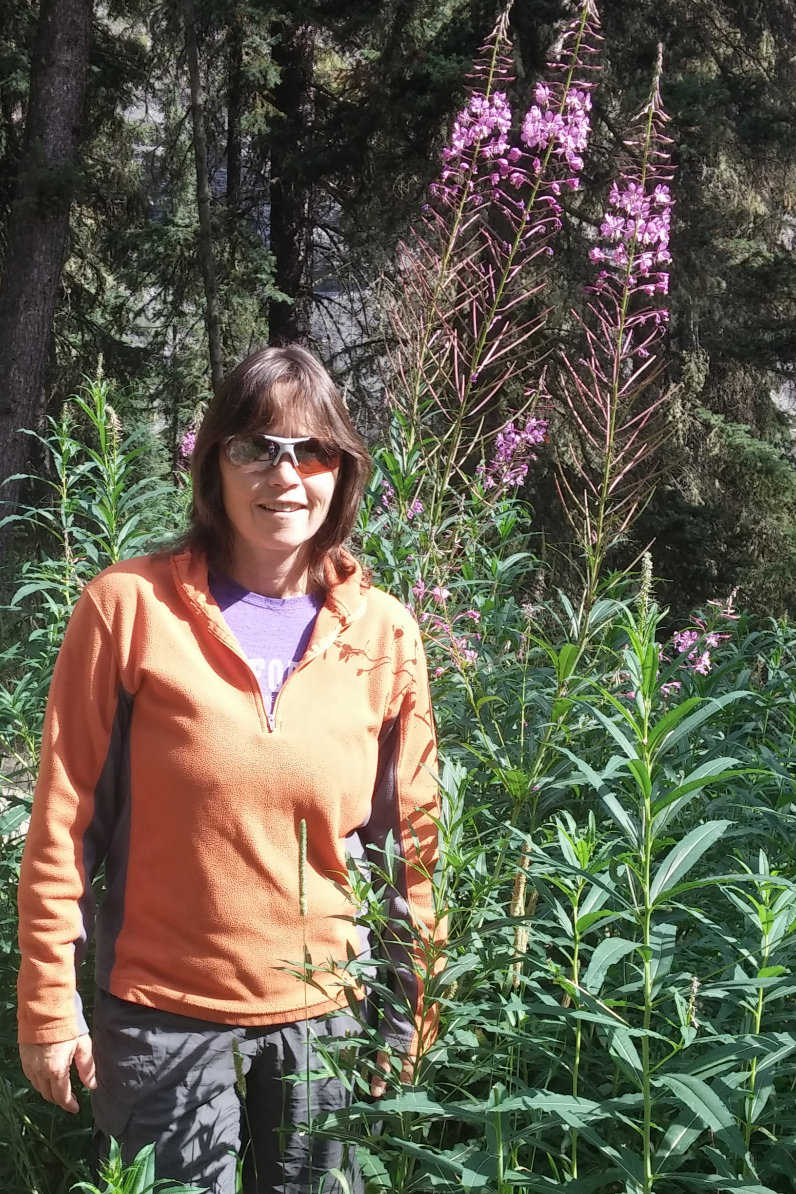 A white woman in an orange fleece and sunglasses stands next to a tall, purple-blooming flower.