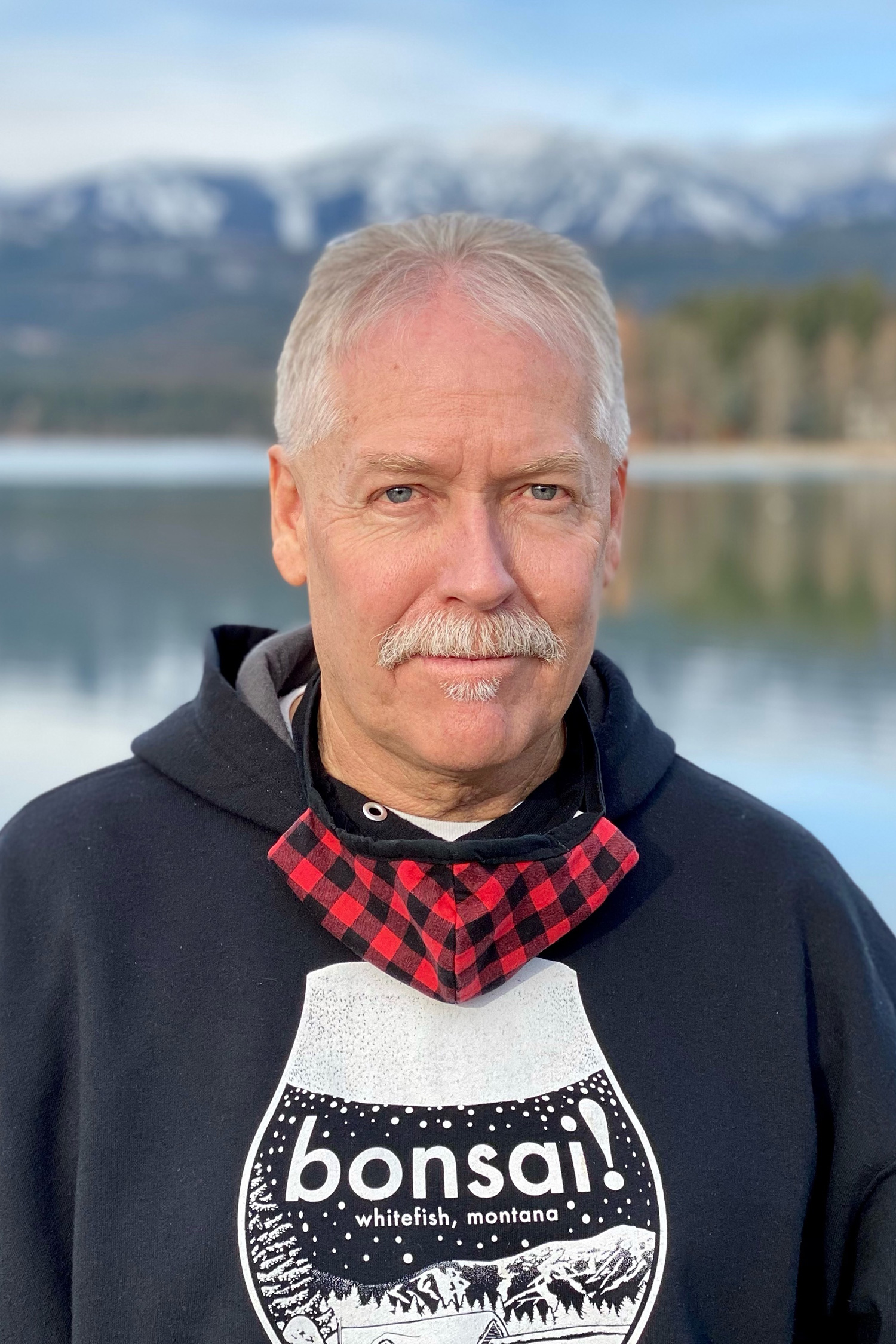 A white man with white hair stands in front of a mountain lake wearing a black hooded sweatshirt.