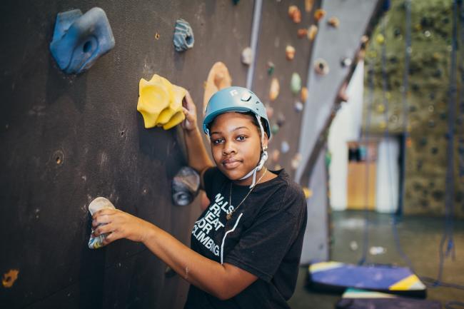A middle school aged girl wearing a climbing helmet prepares to climb an indoor wall