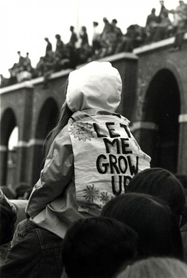 A vintage photo from 1970 of a girl sitting on her parent's shoulders wearing a jacket reading "Let me grow up"