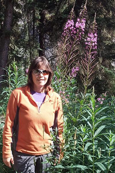 A white woman with brown hair and sunglasses stands in a forest next to tall purple flowers