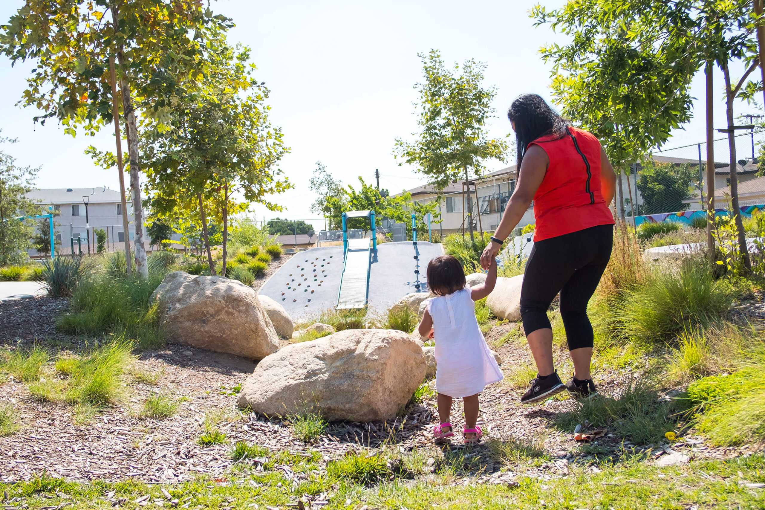 A woman with dark hair in a reddish-orange top leads a young girl in a white dress through a park with trees, a slide, grass, and boulders.