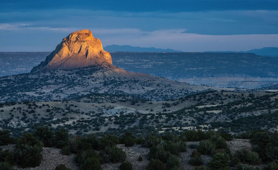 A sunlit peak rises above rolling desert hills covered with shrubs and small trees.
