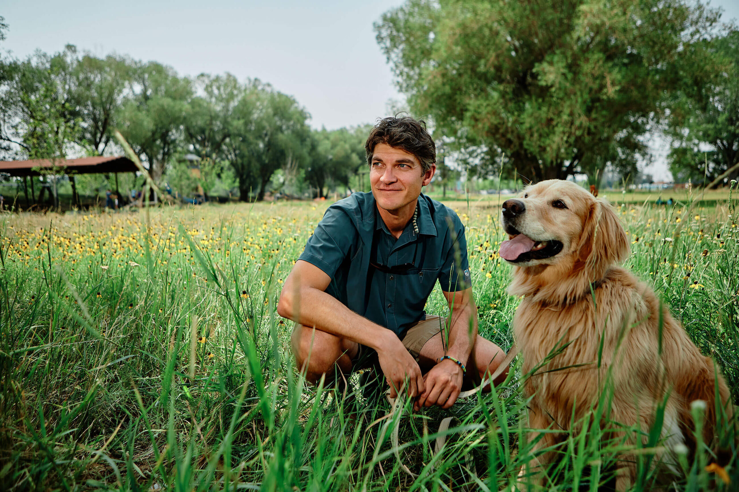 A young man crouching next to a golden retriever in a field.