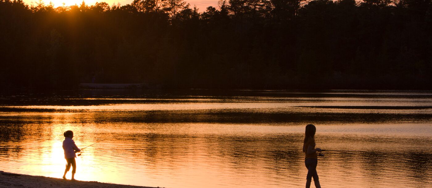 Two people standing on the shore of a lake at sunset.
