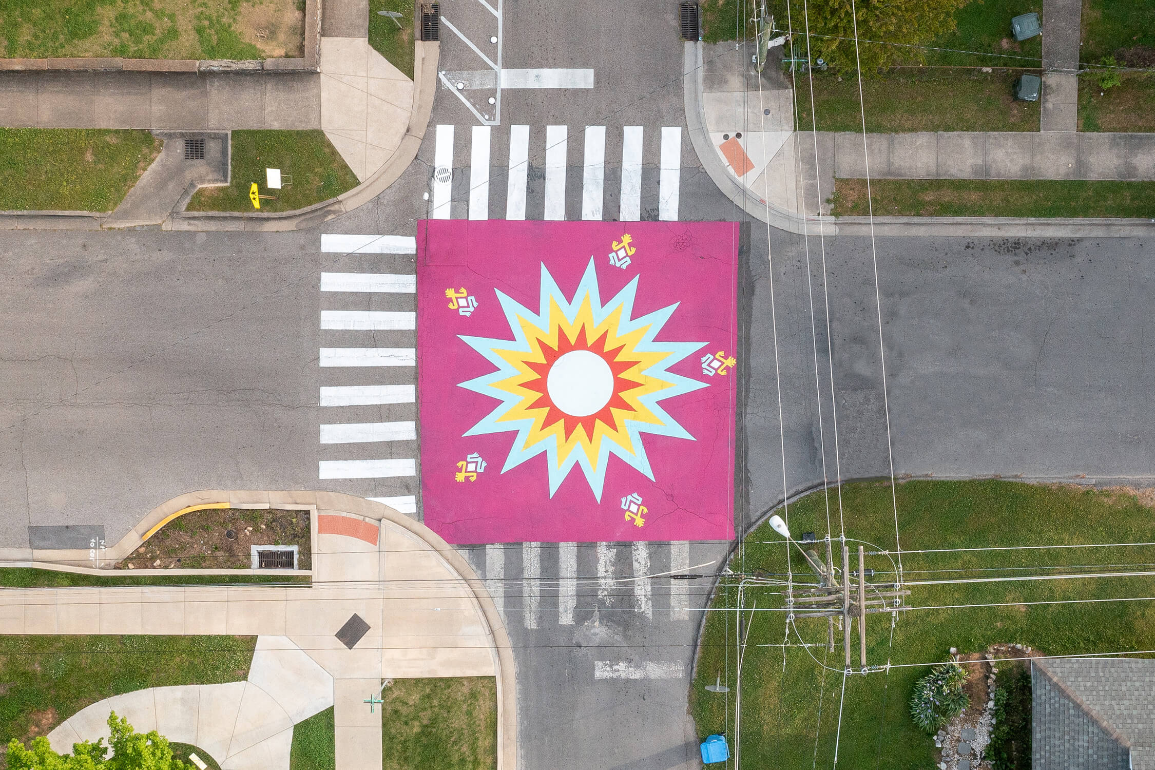A street mural seen from above shows a bright pink square with a starburst in the center and frog designs around the edges.
