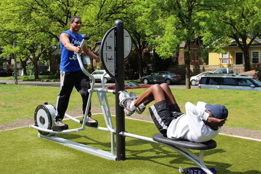 A man and a boy working on an exercise machine in a park.