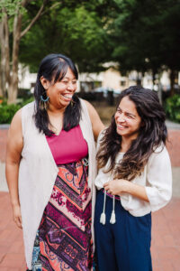 Two dark-haired women stand near a park laughing and smiling at one another.