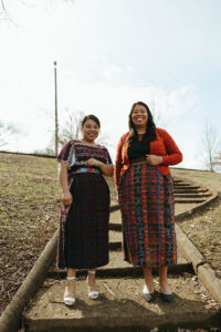 Two dark-haired women in traditional Guatemalan clothing stand on outdoor stairs in a park and smile. 