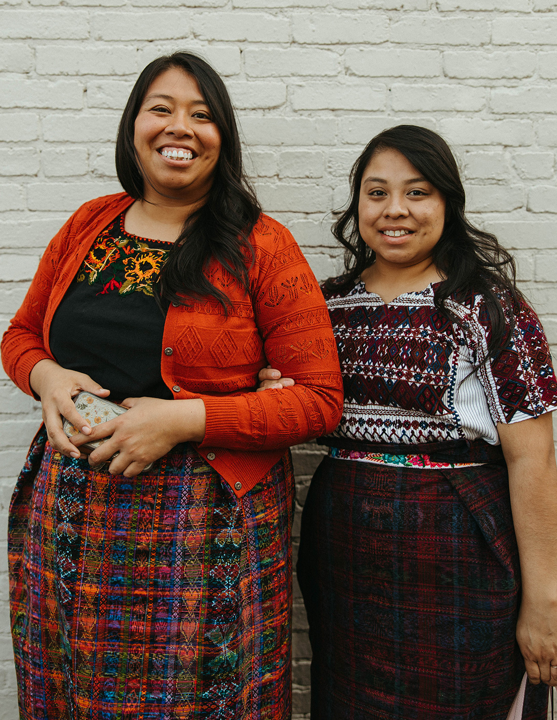 Two dark-haired young women in traditioanl Guatemalan clothing smile and in front of a white brick wall.