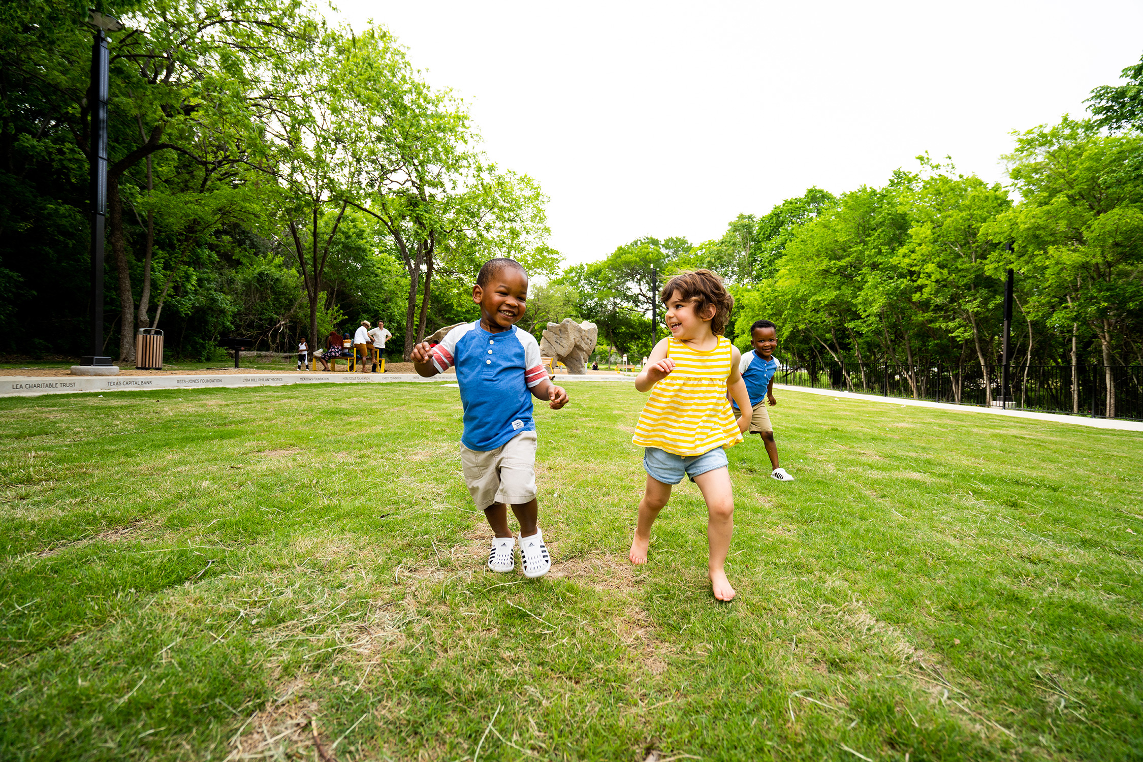 A young boy and girl run across a grassy lawn at a small park edged with trees.