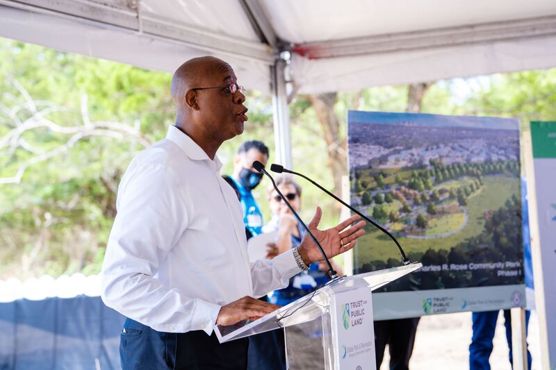 A man stands at a podium and speaks to an audience. There is a rendering a park behind him.