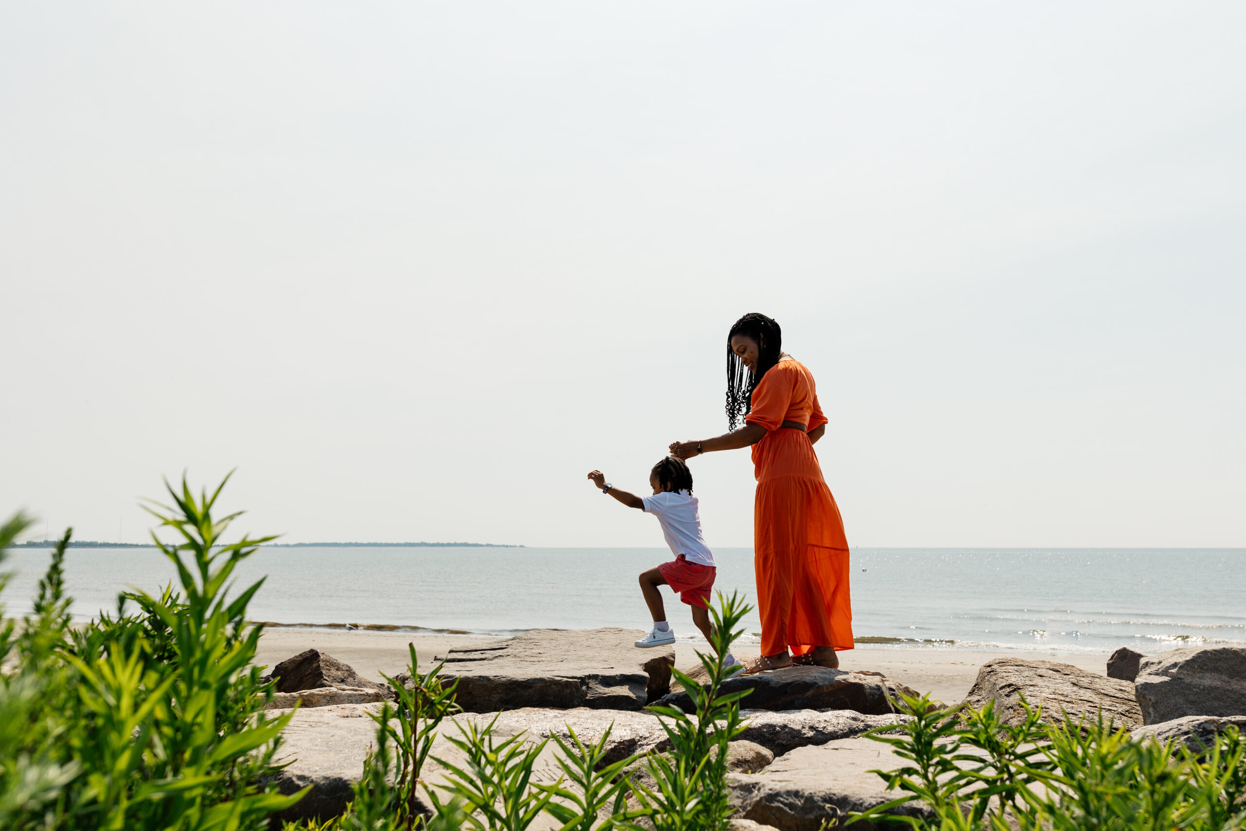 A woman in an orange dress and a child standing on rocks near the ocean.