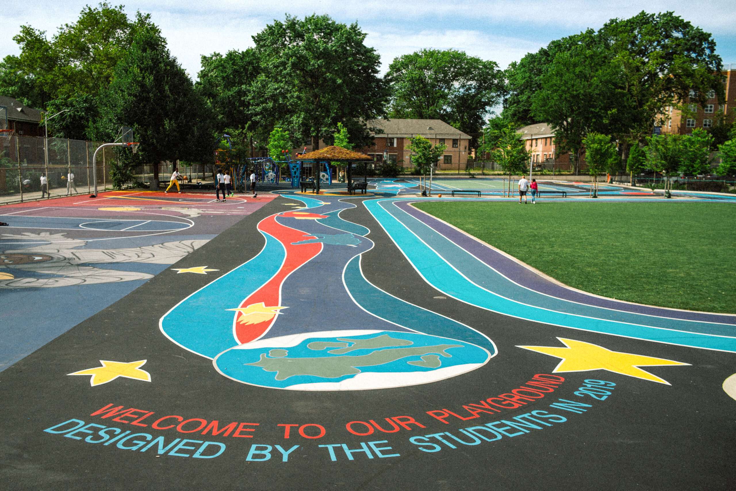 A brightly colored running track surface with the words "Welcome To Our Playground, Designed By the Students in 2019" surrounds a grass playing field at an elementary school.