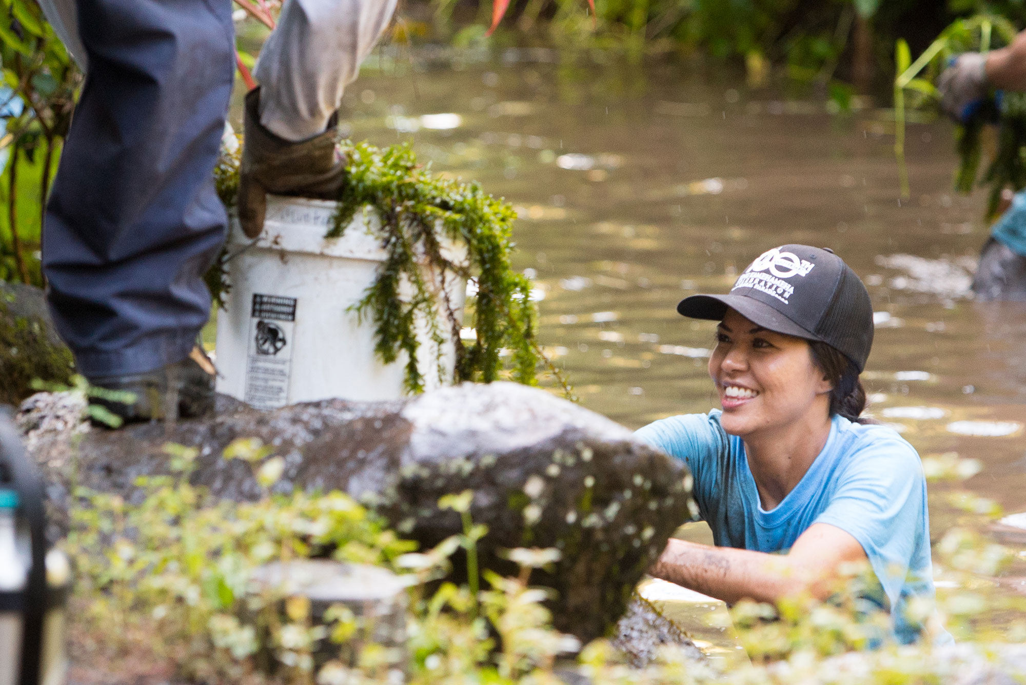 A woman in a hat is standing in a river with a bucket.